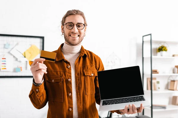 Artista 3d sorridente alla fotocamera e in possesso di carta di credito e laptop in ufficio — Foto stock