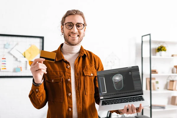 Sorrindo artista 3d olhando para a câmera enquanto segurando cartão de crédito e laptop com projeto de design digital no escritório — Fotografia de Stock