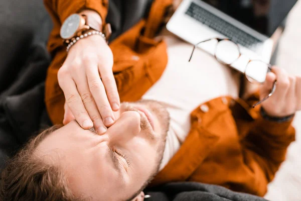 Overhead view of tired 3d designer touching eye and holding eyeglasses with laptop on couch — Stock Photo