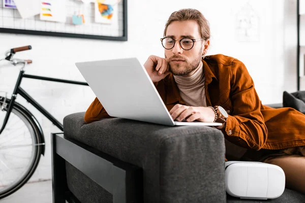 Handsome digital designer working with laptop near vr headset on couch — Stock Photo