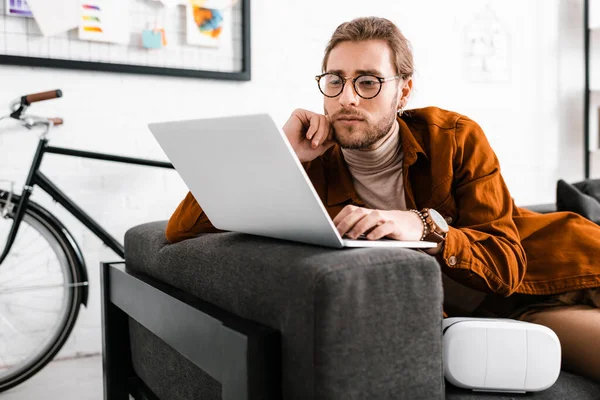 Beau concepteur numérique travaillant avec ordinateur portable près vr casque sur le canapé — Photo de stock