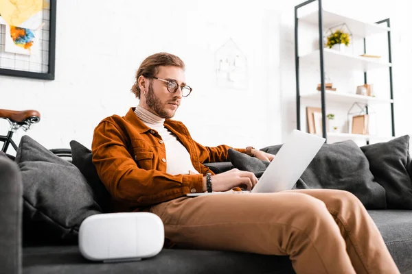 Handsome digital designer using laptop near virtual reality headset on couch in office — Stock Photo