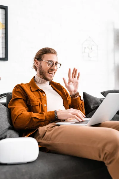 Selective focus of cheerful 3d designer waving hand while having video call on laptop near vr headset on couch — Stock Photo
