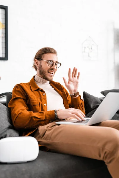 Selektiver Fokus des fröhlichen 3D-Designers, der mit der Hand wedelt, während er Videotelefonie auf dem Laptop in der Nähe des vr-Headsets auf der Couch führt — Stockfoto