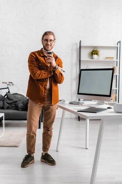 Диджитал-дизайнер улыбается перед камерой, держа кофе рядом с цифровыми устройствами на столе в офисе — стоковое фото