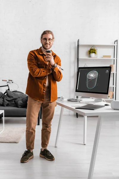 Lächelnder Digitalkünstler blickt in die Kamera, während er Kaffee hält, um in die Nähe von 3D-Design-Projekt auf Computermonitor auf dem Tisch im Büro zu gehen — Stockfoto