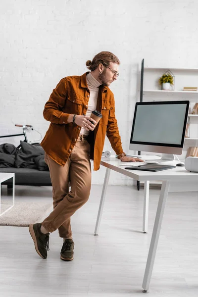 Artista 3d bonito segurando café para ir e olhando para monitor de computador na mesa no escritório — Fotografia de Stock