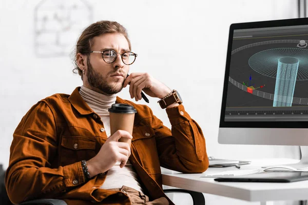 Pensive concepteur numérique tenant café pour aller et stylet près du projet de conception 3D sur écran d'ordinateur sur la table — Photo de stock