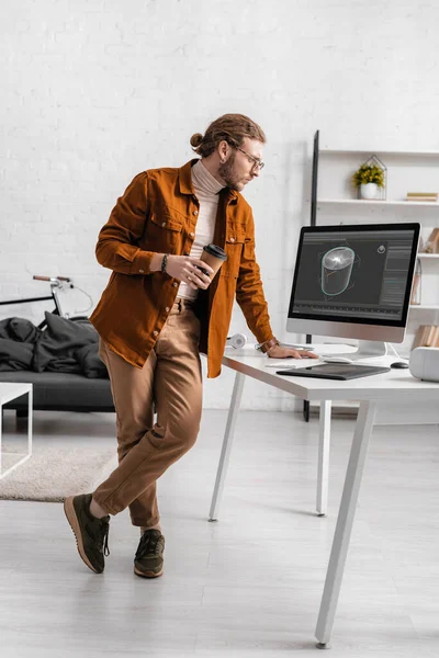 Digitaler Designer hält einen Coffee to go in der Hand und betrachtet ein 3D-Projekt auf einem Computermonitor auf dem Tisch — Stockfoto