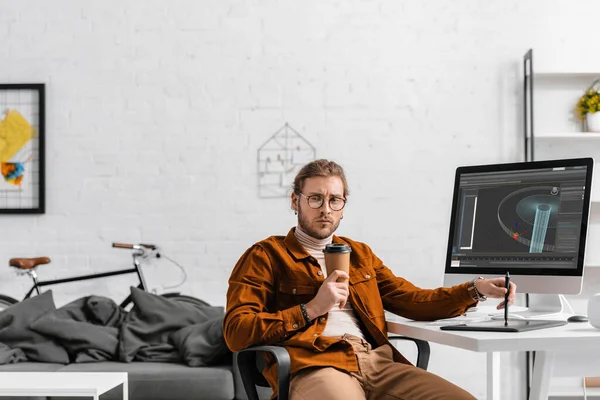 Concepteur numérique réfléchi tenant tasse en papier et stylet près de la tablette graphique et projet de conception 3D sur écran d'ordinateur sur la table — Photo de stock