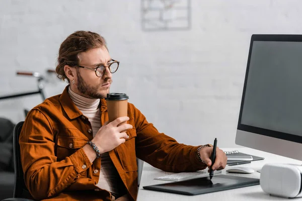 Guapo visualizador 3d celebración de café para llevar y el uso de gráficos tableta cerca de la computadora y auriculares vr en la mesa - foto de stock