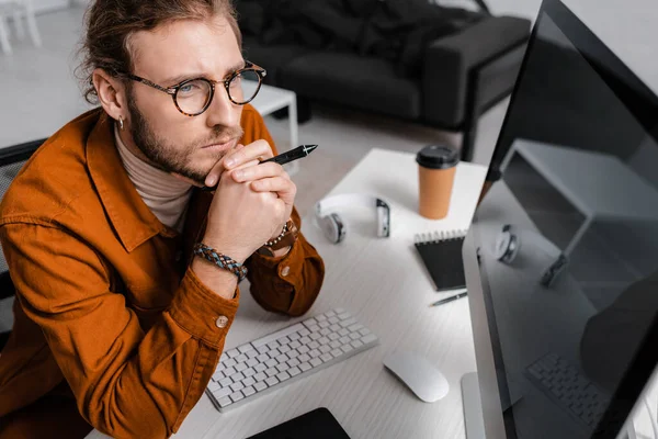 Vista de ángulo alto del visualizador 3D pensativo que sostiene el lápiz de la tableta gráfica cerca de la computadora con la pantalla en blanco en la mesa - foto de stock