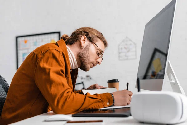 Side view of handsome 3d artist writing on notebook near gadgets on table in office — Stock Photo