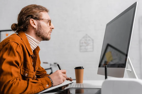 Vue latérale de l'artiste 3d regardant le moniteur d'ordinateur et écrivant sur le carnet à la table — Photo de stock