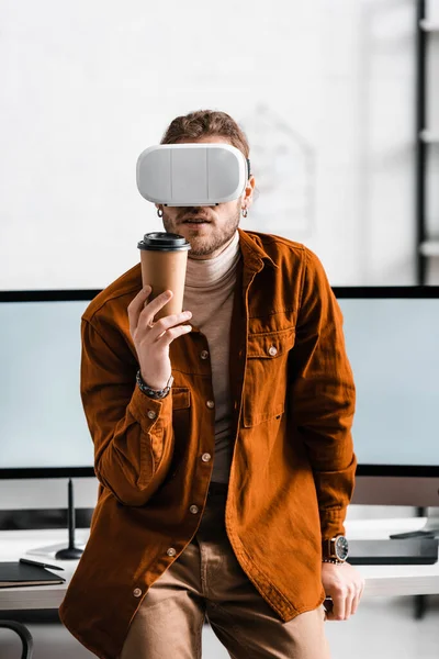 Diseñador digital en auriculares de realidad virtual sosteniendo una taza de papel cerca de monitores de computadora en la mesa - foto de stock
