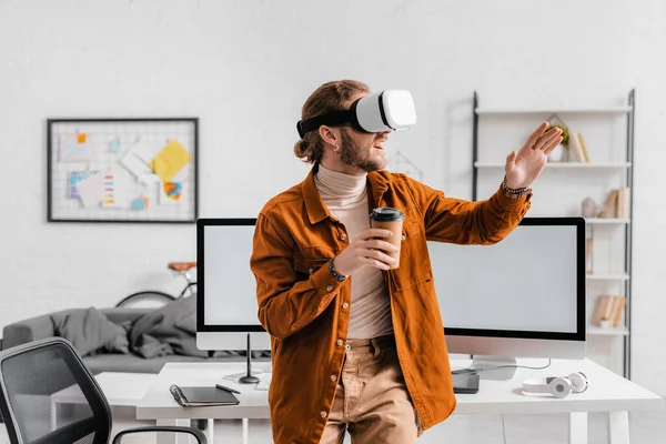 Sonriendo artista 3d utilizando auriculares de realidad virtual y sosteniendo el café para ir en la oficina - foto de stock