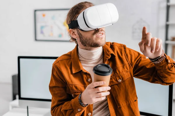 3D художник в наушниках виртуальной реальности держит кофе, чтобы пойти и показывать пальцем в офисе — стоковое фото
