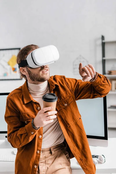 Digitaldesigner zeigt mit dem Finger auf Virtual-Reality-Headset und hält Pappbecher im Büro — Stockfoto