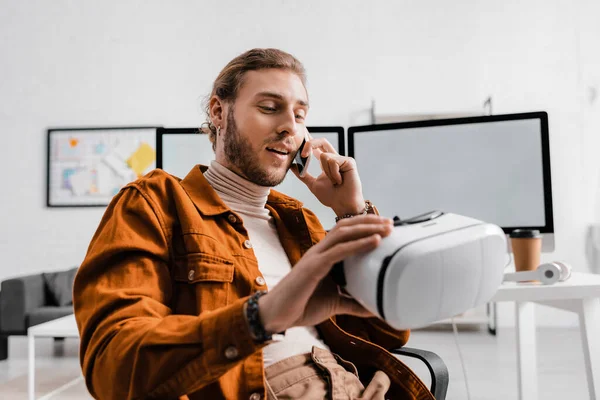Enfoque selectivo del guapo diseñador digital que habla en el teléfono inteligente y sostiene los auriculares de realidad virtual en la oficina - foto de stock