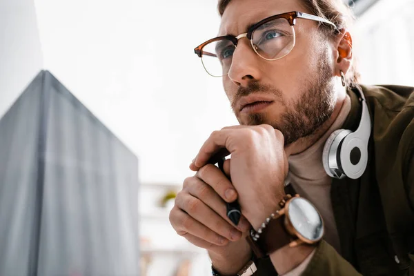 Pensive 3d artist in headphones holding stylus of graphics tablet near computer monitor with black screen in office — Stock Photo