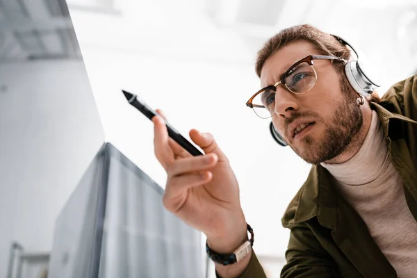 Вид з низьким кутом 3d художника в навушниках, що тримає стилус графічного планшета біля монітора комп'ютера з чорним екраном в офісі — стокове фото