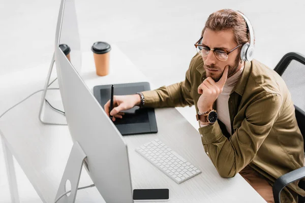 Hochwinkelaufnahme des digitalen Designers in Kopfhörern mit Grafik-Tablet und Computer am Tisch auf grauem Hintergrund — Stockfoto