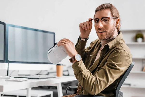 Handsome 3d artist holding vr headset and looking at camera at table in office — Stock Photo