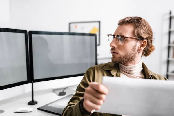 Selective focus of handsome digital designer holding blueprint and looking at computer monitors on table — Stock Photo