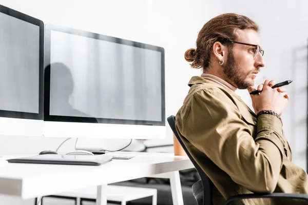 Side view of handsome 3d artist holding stylus of graphics tablet and looking away near computers on table — Stock Photo