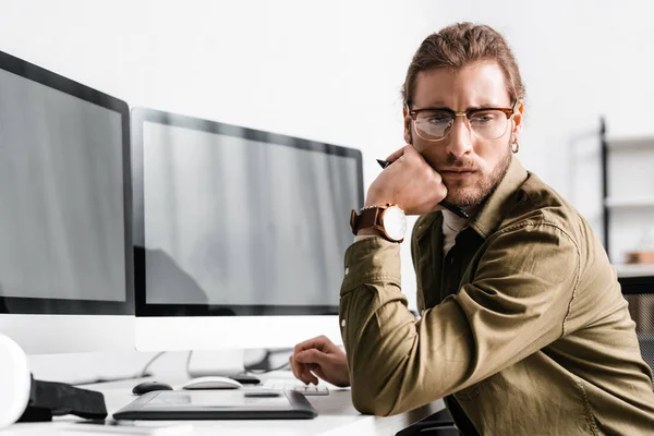 Diseñador digital pensativo mirando hacia otro lado cerca de gráficos tableta y ordenadores en la mesa - foto de stock