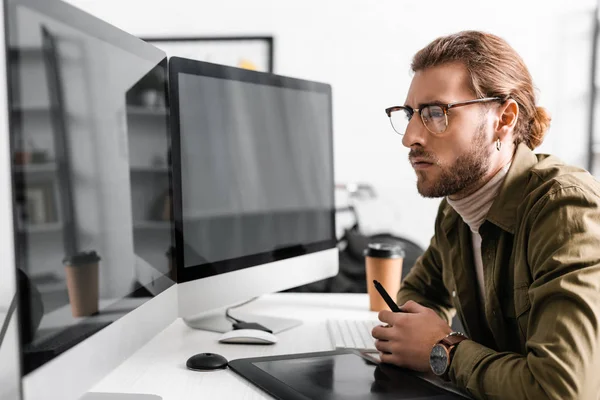 Вид збоку гарний 3d художник тримає стилус біля графічного планшета і дивиться на комп'ютерні монітори з порожнім екраном на столі — стокове фото