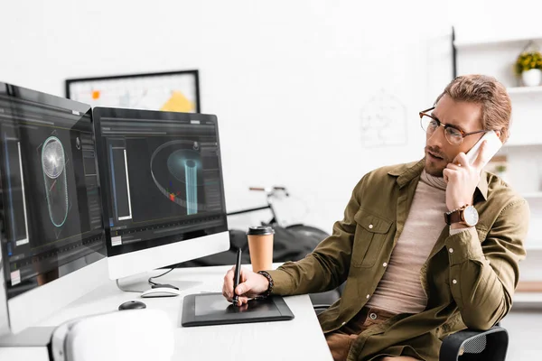 3D художник разговаривает на смартфоне во время творческого проекта 3D дизайна на компьютерах за столом в офисе — стоковое фото