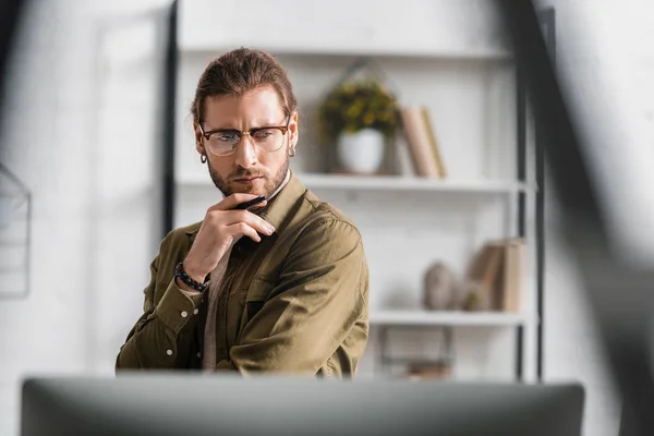 Selective focus of pensive digital designer holding stylus of graphic tablet and looking at computer monitor in office — Stock Photo