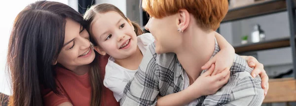 Panorámica de la sonriente pareja del mismo sexo abrazando a su hija en casa - foto de stock