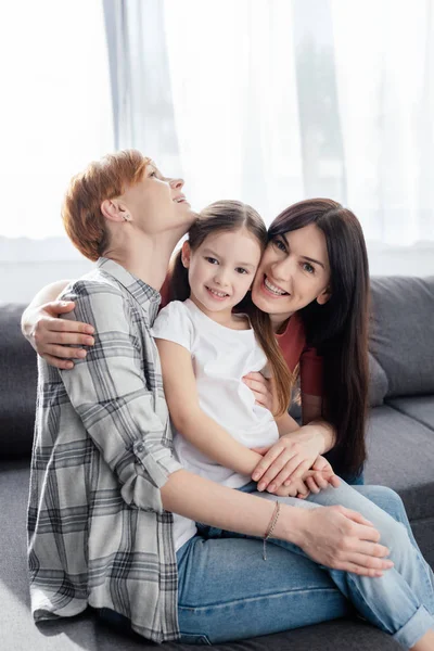 Fröhliche gleichgeschlechtliche Eltern umarmen Tochter und blicken auf Couch im Wohnzimmer in die Kamera — Stockfoto