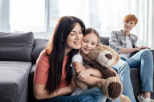 Selektiver Fokus einer lächelnden Mutter, die Tochter mit Teddybär in der Nähe eines Elternteils auf der Couch im Wohnzimmer umarmt — Stockfoto