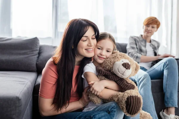 Enfoque selectivo de la madre sonriente abrazando a la hija con el oso de peluche cerca de los padres en el sofá en la sala de estar - foto de stock