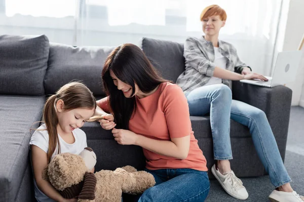 Foco seletivo de mãe sorridente perto da filha com ursinho de pelúcia e pai com laptop no sofá em casa — Fotografia de Stock