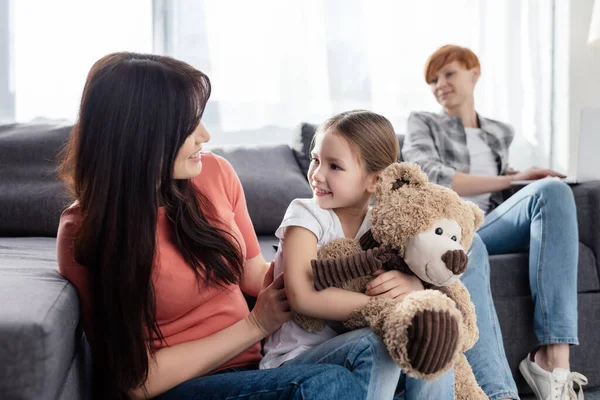 Selektiver Fokus des Kindes mit Teddybär, das Mutter nahe Elternteil mit Laptop auf der heimischen Couch anlächelt — Stockfoto