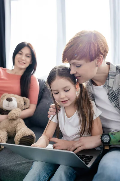 Selective focus of woman hugging daughter while using laptop near parent with teddy bear on couch — Stock Photo