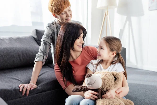 Sonrientes padres del mismo sexo mirando a su hija con oso de peluche en la sala de estar - foto de stock