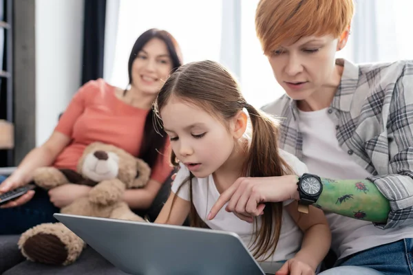 Enfoque selectivo de la madre usando el ordenador portátil con la hija cerca de padres sonrientes con oso de peluche en el sofá - foto de stock