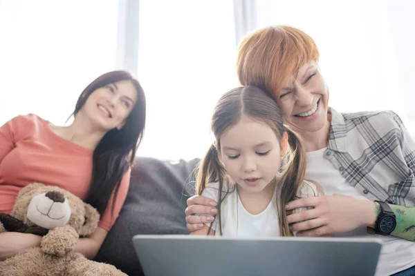 Focus selettivo di madre allegra abbracciando figlia con computer portatile vicino genitore sorridente con orsacchiotto sul divano — Foto stock