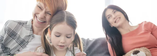 Selective focus of cheerful woman hugging daughter near smiling parent with teddy bear on couch, panoramic shot — Stock Photo