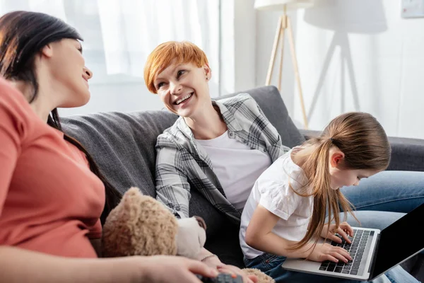 Selektiver Fokus eines lächelnden gleichgeschlechtlichen Paares, das sich in der Nähe eines Kindes mit Laptop auf der Couch anschaut — Stockfoto