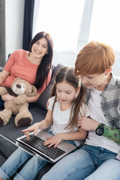 Выборочный фокус матери с помощью ноутбука с дочерью рядом улыбающийся родитель с плюшевым мишкой на диване дома — стоковое фото