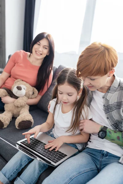 Selective focus of mother using laptop with daughter near smiling parent with teddy bear on couch at home — Stock Photo