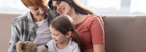 Panoramic shot of smiling same sex couple sitting near daughter with teddy bear on couch — Stock Photo