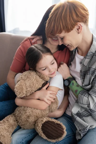 Glückliche gleichgeschlechtliche Eltern umarmen Tochter mit Teddybär auf Couch — Stockfoto