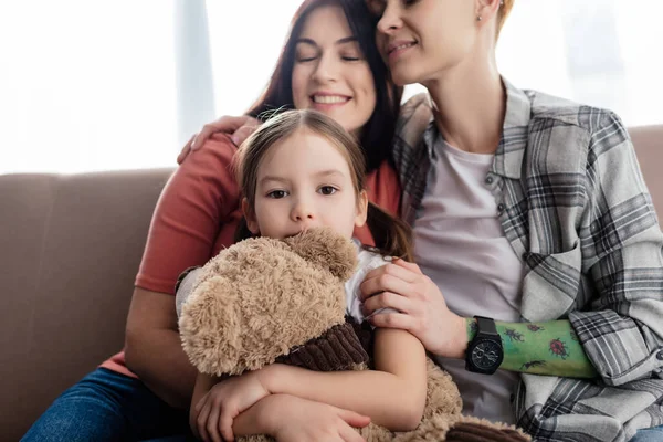 Child with teddy bear looking at camera near smiling same sex parents on couch — Stock Photo
